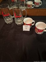 View of two Slim Jim glass mugs with red logo and two Campbell Soup collectible ceramic mugs sitting on black surface with packaging.