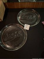 Two round heavyweight glass serving trays with embossed Coca-Cola logo, shown on a dark surface with lot number tag.