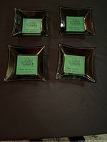 Four black reflective glass square trays arranged in a 2x2 grid showing green labels with ship illustration and 'Hotel Mayflower New York City' text.