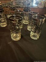 Photo showing multiple clear glass tumblers with a decorative script 'C' on each glass, arranged on a dark surface.