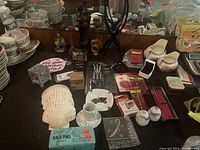 Photo showing assortment of household items including plates, large seashells, boxed Diane hair pins, salt and pepper shakers, a universal remote, and a wig stand for sale.
