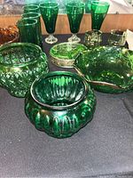 Photo showing the full set of vintage forest green glass items on a black surface, including glasses, planter vase, bowl, tray holder, and dish.