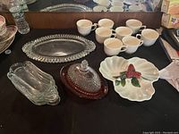 Overview of most items in lot including glass serving tray with raised edge, eight white ceramic cups, clear divided glass tray, candy dish with lid, leaf-shaped ceramic dish with red berries and green leaves.