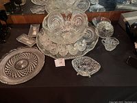 Two sets of crystal punch bowls with accompanying matching cups, various serving trays and dishes on a black surface with a mirrored background.
