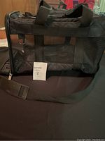 Black small pet travel bag with mesh ventilation panels, black handles, and adjustable shoulder strap. The bag is placed on a dark surface and photographed from the side showing details.