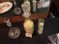 Three glass items including tall faceted bottle with stopper filled with yellow bath salts, tall narrow container with layered salts/soap, and rounded decanter with stopper.