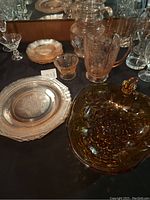 Photo of vintage glassware set on black cloth featuring pink pitchers, plates, small cup, and amber grape serving bowl
