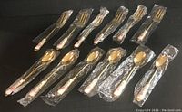Twelve pieces of cutlery: six small forks and six small spoons wrapped in original plastic packaging, arranged on a black surface.