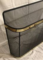 Close-up image showing black mesh screen with shiny brass trim and one brass handle mounted at the center top edge, highlighting mesh texture and brass finish.