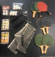 Overview photo showing 7 ping pong paddles, black mesh net, metal clamps and multiple packs of white balls