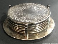 Stacked set of six silver-plated coasters in round matching holder showing top engraved surface and holder base with four supporting feet