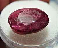 Close-up image of the oval faceted ruby gemstone on white foam background showing surface details and inclusions.