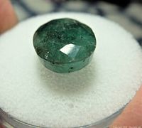Close-up photo showing the emerald gemstone's round faceted shape, deep green color, and internal inclusions.