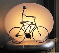 Table lamp featuring a lit white circular shade and a silver metal bicyclist sculpture on a bicycle mounted on a silver base with a solar power plaque.