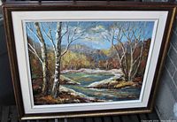 Framed signed oil painting on board depicting a river landscape with trees and mountains, photographed from front with signature visible.