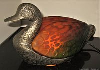 Close-up of the lamp showing silver metal duck head and glowing art glass body.