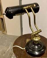 Photo of black and brass adjustable banker’s desk lamp on a wooden stool, showing the lamp base, arm, and cylindrical shade with ventilation holes.