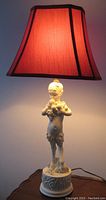 Full view of porcelain satyr themed table lamp with red fabric trapezoidal shade lit in a dim room.