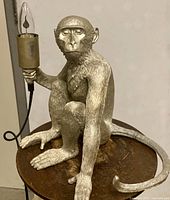 Front view of silvered monkey table lamp holding a flame-shaped bulb, sitting on a round base. Cord visible.