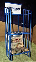 Blue metal newspaper box stand with two shelves and Toronto Star logo sign on top with five miniature newspapers on bottom shelf.