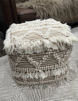 Woven textured wool and jute pouf with fringe on edges and geometric patterns in neutral tones, providing visual detail and condition.