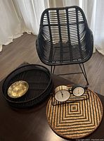 Black rattan accent chair with metal legs, two round rattan trays, small metal bowl, and a dual dial clock arranged on wooden floor.