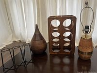 Three items standing in front of a white curtain: wooden vase, wooden bamboo wine bottle holder, wooden lamp base with metal harp.