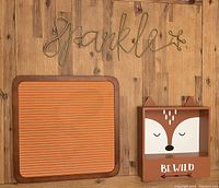 Three wall decor items displayed on a wooden floor: gold metal 'Sparkle' slogan, striped board, and fox face shelf with 'Be Wild' text.