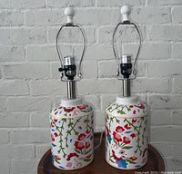 Front view of both floral ceramic lamps with attached harp frames on wood table against white brick wall.