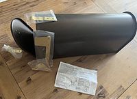 Photo showing the bronze mailbox body, several gold-tone mounting brackets in bags, a doorstop or spacer, and paper installation instructions on a wooden floor.