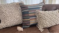 Photo showing three throw pillows/covers on a brown leather couch: a textured beige pillow, a striped multicolor square pillow cover with tassels, and a beige macrame rectangular pillow cover with tassels.