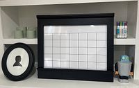 Photo showing whiteboard calendar with grid layout in black frame, dry erase markers on shelf, black oval frame and robot coin bank on shelf
