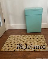 Light blue sensor trash can and Francesca Daisy Welcome door mat placed on floor against wall