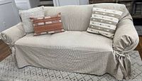 Full sofa covered with beige textured washable fabric cover tied at the sides and four decorative throw pillows of various colors and sizes.