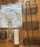 3 tier black metal wall mount organizer with three wire baskets, white cylindrical water filter with tubing and faucet, small black wire basket, clear plastic component with metal base, and fabric organizer laid out on floor.