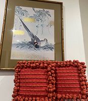 Photo showing framed bird print hung on wall above two red throw pillows with pom-pom trim on couch.