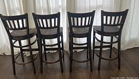 Four wooden bar stools shown from rear view displaying ladder-back design and circular foot guards.