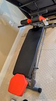 Full view of abdominal bench showing black padded main surface with orange headrest and leg pads, metal frame visible.
