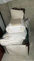 Stack of bedding items including comforters in protective fabric bags and folded sheets and pillows on a cement floor.