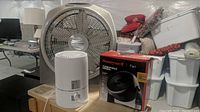 Three air circulation products on a wooden table in a basement setup: Honeywell Turbo Force fan box, white air purifier, and Lasko Wind Tunnel fan