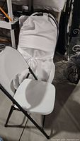 Front angle view of one white plastic folding chair with decorative white satin-like cover tied with a bow on the backrest.