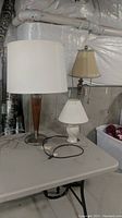 Shows three lamps on a grey folding table in a basement; ceramic table lamp, wood and chrome table lamp, brass look resin floor lamp