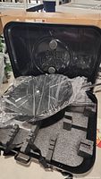 Black plastic prize wheel inside a large black molded carrying case with foam inserts; wheel is wrapped in plastic and comes with a detachable stand.