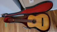 Goya classical guitar in open black hard shell case with red plush interior; silver harmonica visible near guitar body