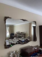 Large wall mirror with ornate bronze-colored decorative corner accents hung on a beige wall, showing reflection of a dining area and living room.