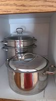 Stainless steel pots in cabinet, showing a double steamer pot stacked and a large pot with lid in front