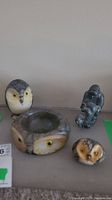 Four stone carvings displayed together on a shelf against a neutral background. Includes three alabaster owl themed pieces and one dark gray Indigenous soapstone carving.