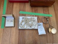 Wide view of all items included in the lot on floor: earrings in box, pearl necklace in box, gold-tone locket necklace, and Timex watch