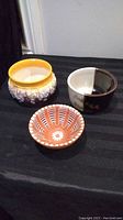 Three ceramic bowls placed on a black cloth; front bowl has orange and white radial design, other two include a yellow rim drip glaze bowl and a black and white floral two-tone bowl.