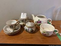 Two teacups with seven saucers, sugar bowl, matching floral decorations, and teapot with separate cup/lid shown on wooden surface